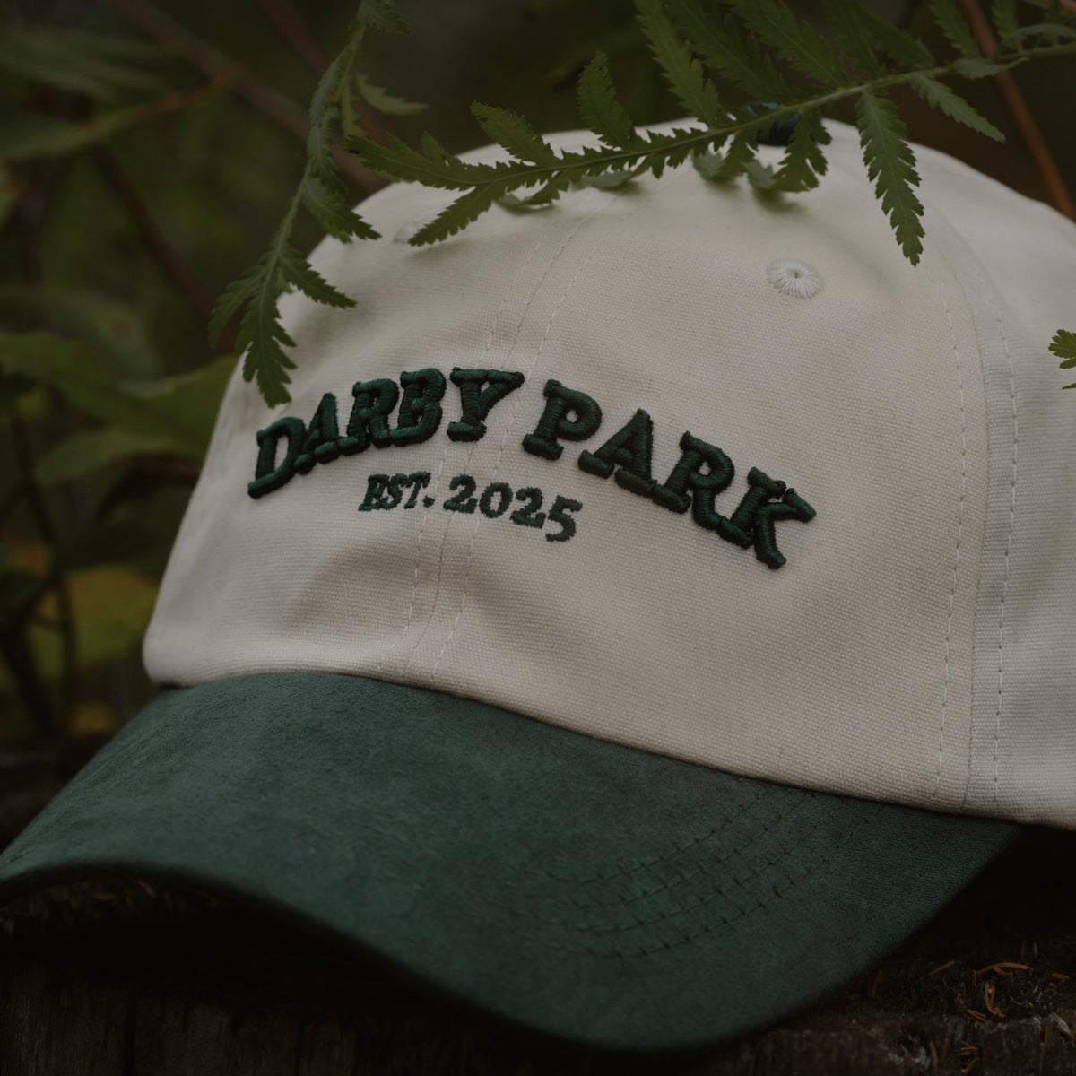 The Canvas Cap in cream cotton canvas with a green faux suede brim and embroidered Darby Park wordmark, under pine branches.