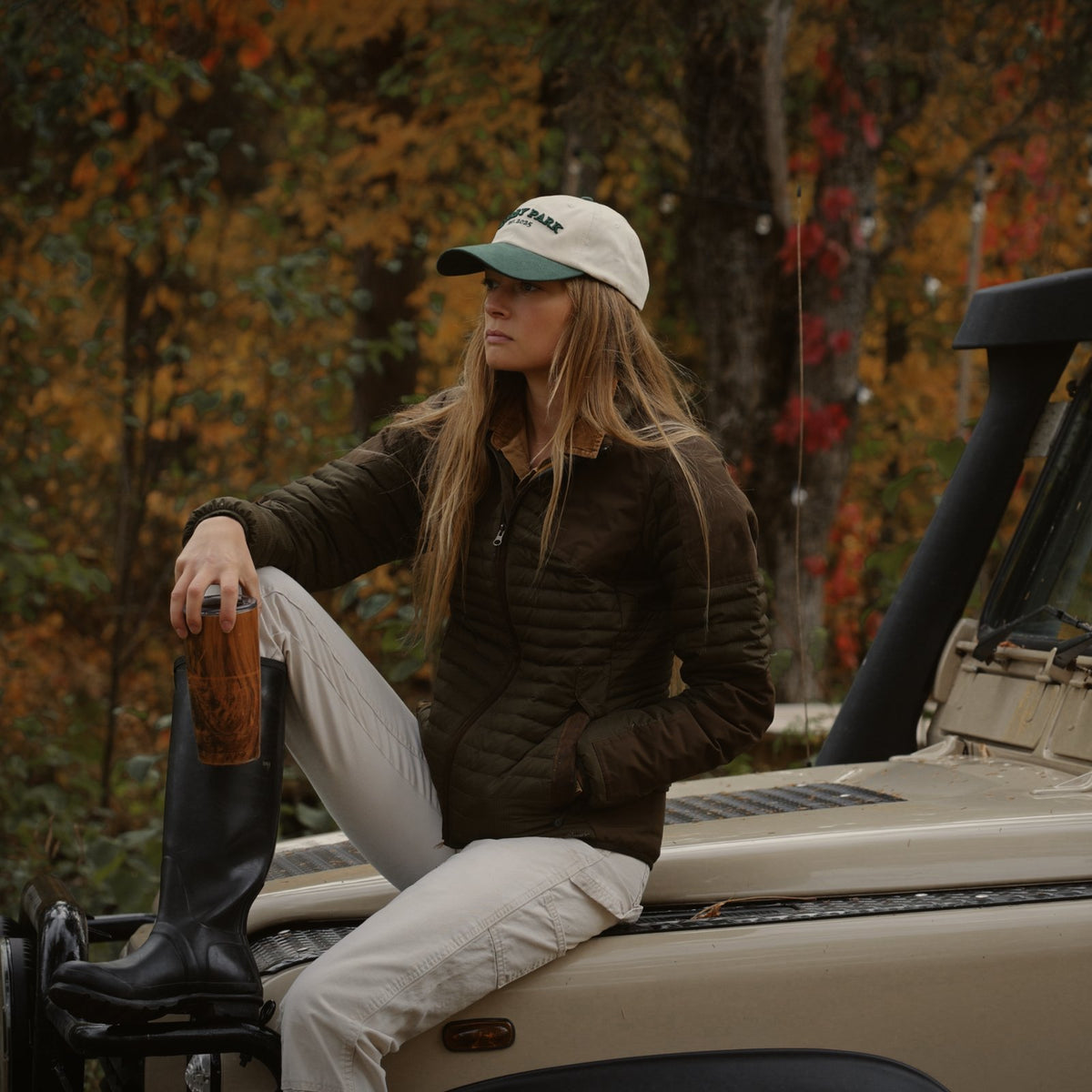 Woman in the Canvas Cap and a quilted jacket sitting on the hood of a vintage truck in the autumn woods.