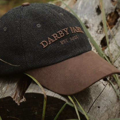 The Tweed Cap in dark green wool-blend herringbone with a brown faux suede brim and embroidered Darby Park wordmark, resting on a weathered log.