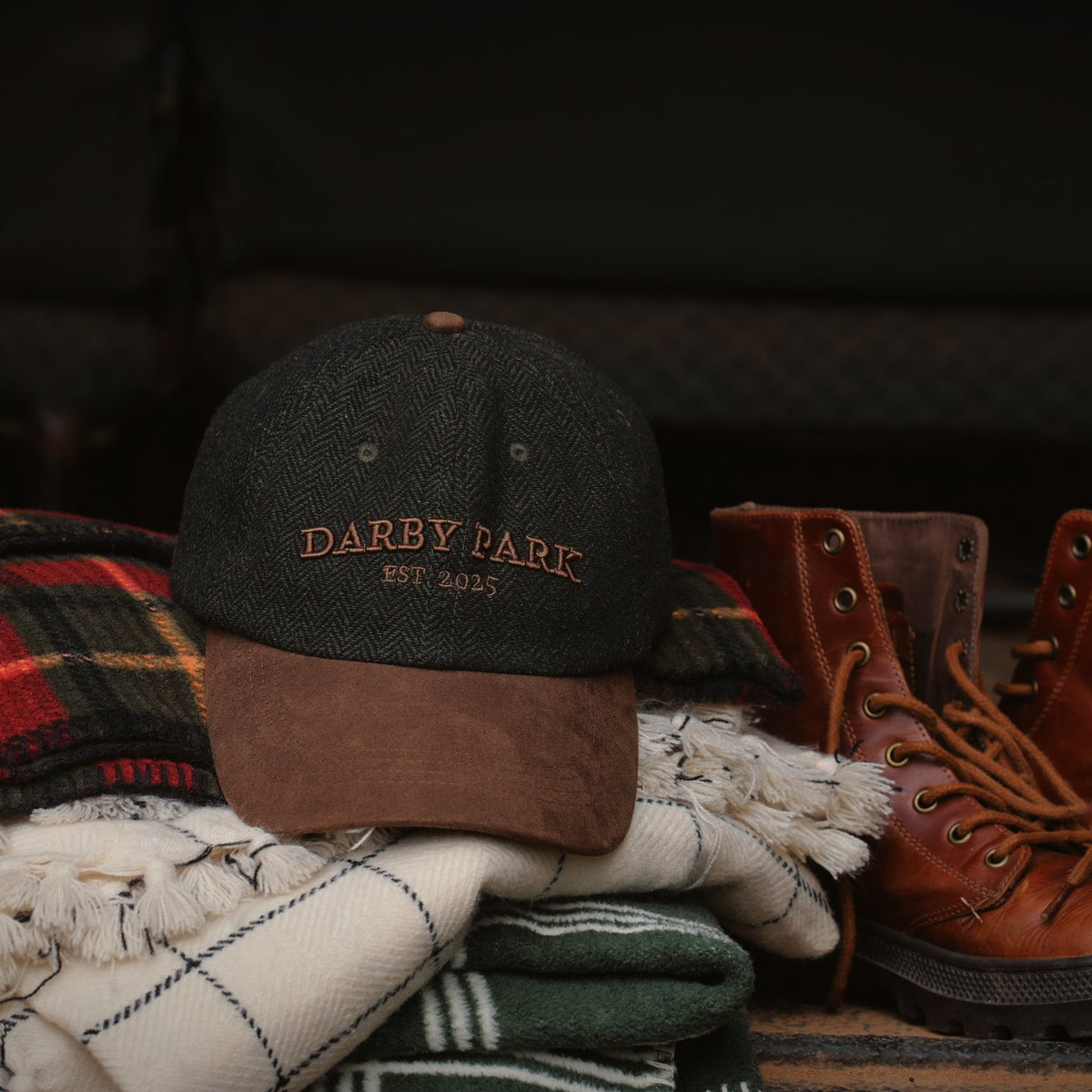 The Tweed Cap on a stack of folded wool blankets beside leather boots.