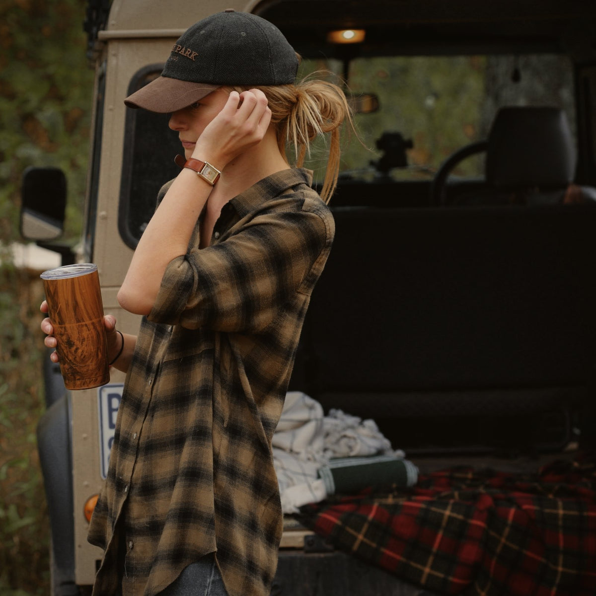 Woman in the Tweed Cap and a plaid flannel shirt holding a coffee beside an old truck.
